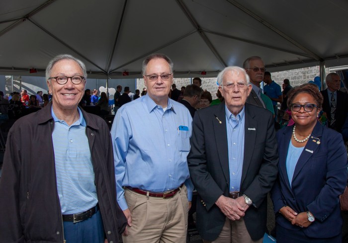 under a white tent outdoors, 3 men and 1 woman pose for a group photo. 2 of the individuals wear a shovel-shaped pin