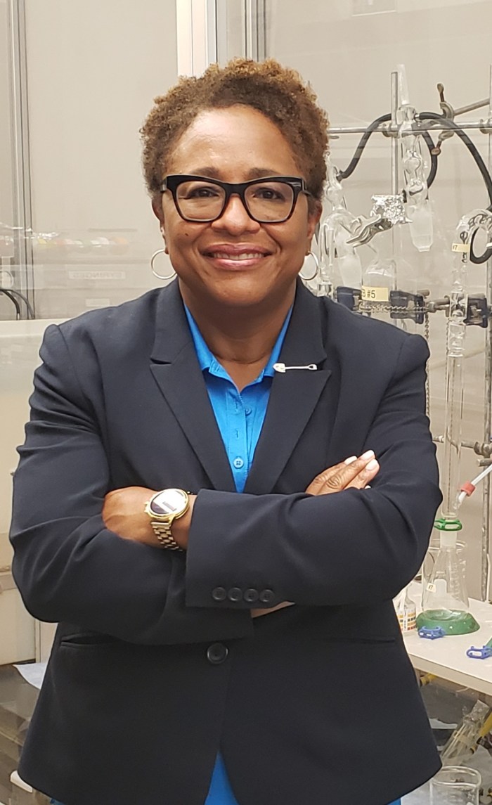 woman utility supervisor with short brown curly hair and glasses, blue collared blouse, and navy blue blazer with shovel-shaped pin on her lapel posed for photo as she stands in front of a tall rack of lab equipment and clear glass beakers