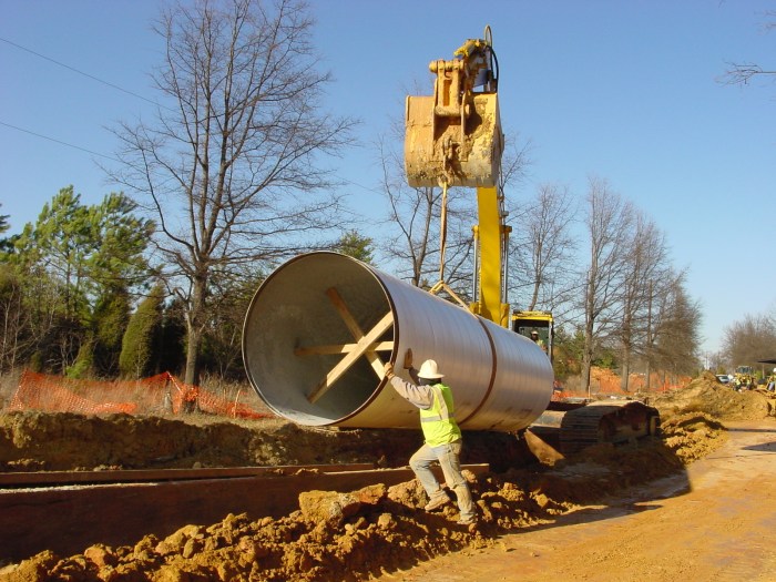 large yellow excavator with heavy gauge rope attached to its bucket lowers a very large section of white pipe into a trench in the ground, nearby a utility technician wearing a yellow safety vest and white hard hat stands on recently dug rocks guiding the pipe into place by pushing on it