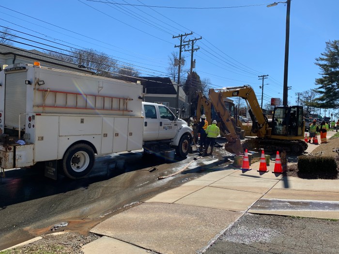 on a city street, a large white utility truck parks near a job site, nearby are 3 orange traffic cones, 2 utility technicians in orange and yellow safety vests and white hard hats and 2 yellow backhoes, a tan hose is unfurled along the gutterway