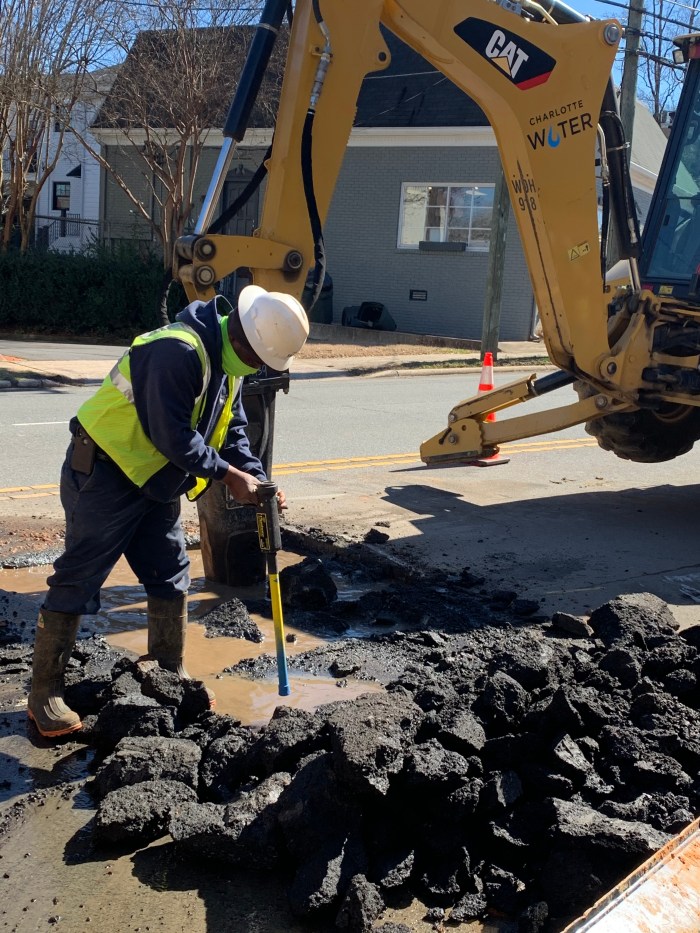 on a city street, a utility technician wearing blue coveralls, yellow safety vest and white hard hat use a hand drill with yellow and blue extender to help unearth something below a pool of muddy water in the rectangular pit recently dug by the yellow CAT backhoe behind him