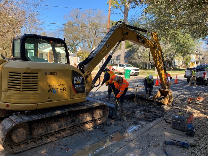 on a suburban tree-lined street, 3 utility technicians wearing orange and yellow safety vests and white hard hats dig dirt with shovels as they stand in a square pit cut out by a nearby CAT, backhoe, in the background are 2 white utility pickup trucks and a fourth utility technician