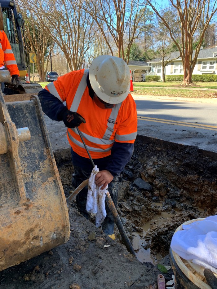 utility technician wearing orange safety shirt and white hard hat uses a brown pipe shaped tool to hand-pump his way into a section of a rectangular dirt pit cut into the side of a suburban tree-lined street