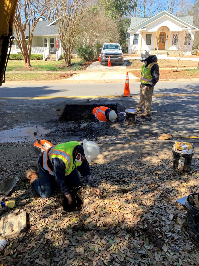 wearing orange and yellow safety vests and white hard hats, 2 utility technicians dig curbside along a suburban street while a third stands by an orange traffic cone looking down into the larger square hole containing one of the technicians