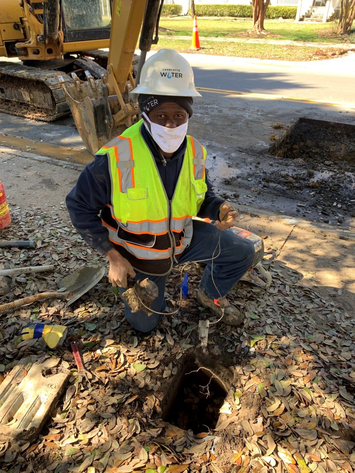 curbside on a suburban street, next to a yellow backhoe, a utility technician in blue coveralls, yellow safety vest and white hard hat kneels on brown-bark littered ground next to a removed rectangular white water meter lid revealing a rectangular hole in the ground
