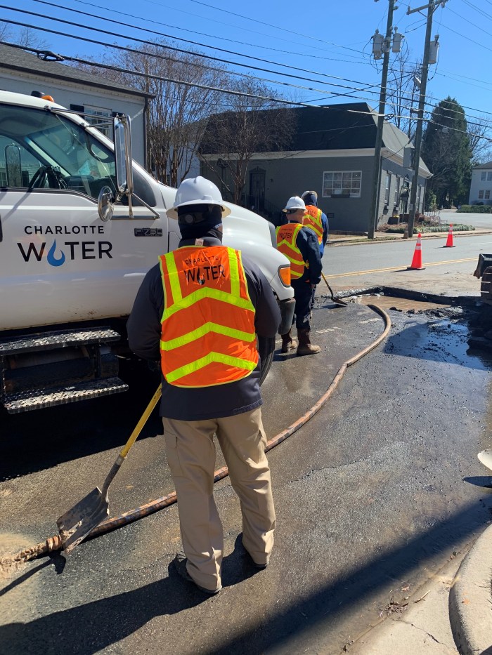 utility technician wearing orange safety vest and white hard hat stands on wet street with shovel as a tan hose runs by underneath him, next to him is white utility truck with Charlotte Water logo and a shallow cut square pit and two other utility technicians in orange safety vests along with 2 orange traffic cones