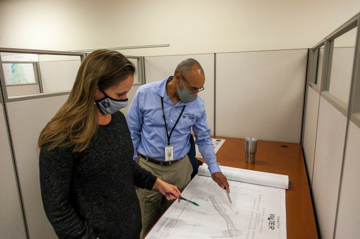 2 engineers wearing office clothes stand in an office cubicle looking down at a large white, unrolled schematic on a brown desk, both engineers point at the image of a waterway on the schematic with their pens