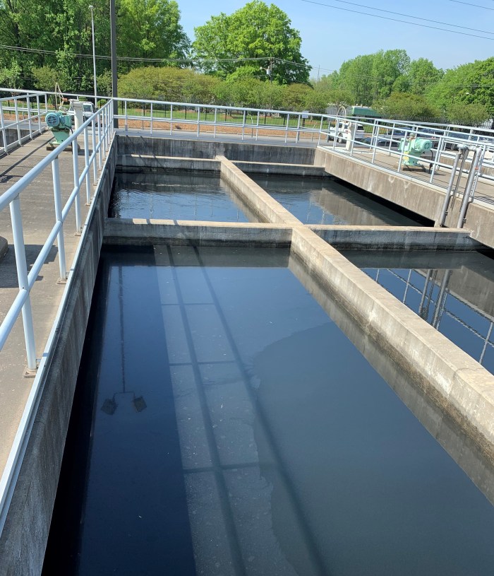 surrounded by metal fencing at a water treatment facility, an outdoor rectangular concrete pond has four symmetrical sedimentation chambers