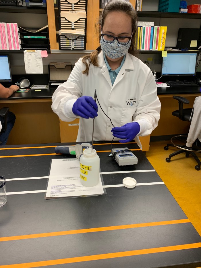 standing a at black table, a lab technician wearing floral printed mask, glasses, white lab coat and blue gloves inserts a testing probe into an open sample jar, probe is attached to a nearby small, grey plastic meter with a black wire
