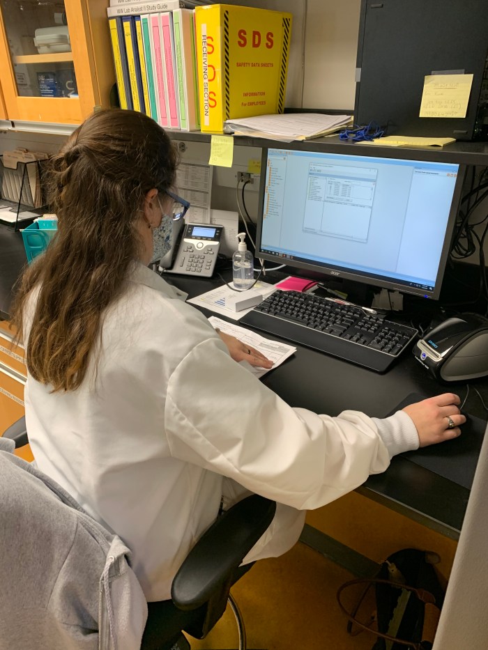 lab technician wearing glasses, floral printed mask and white lab coat sits at desk looking a computer screen