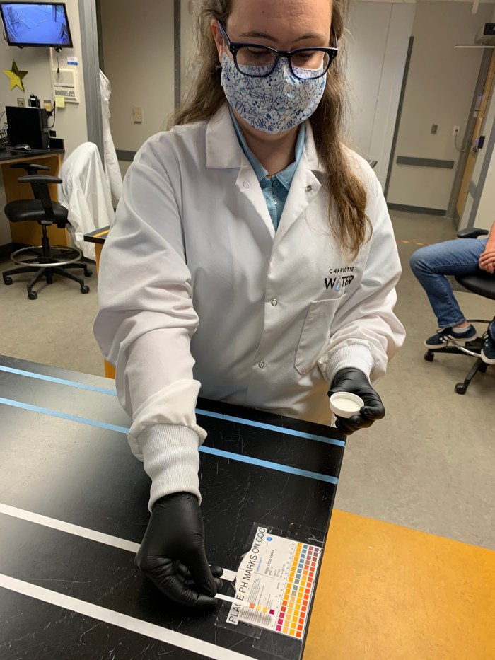 lab technician with glasses, floral print mask, white lab jacket, black gloved hands holds a colorful ph lab strip against a ph chart as comparison