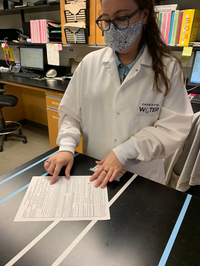 lab technician wearing glasses, floral print mask, and white lab coat with Charlotte Water logo, stands at black lab table pointing at a section of text on a printed piece of paper