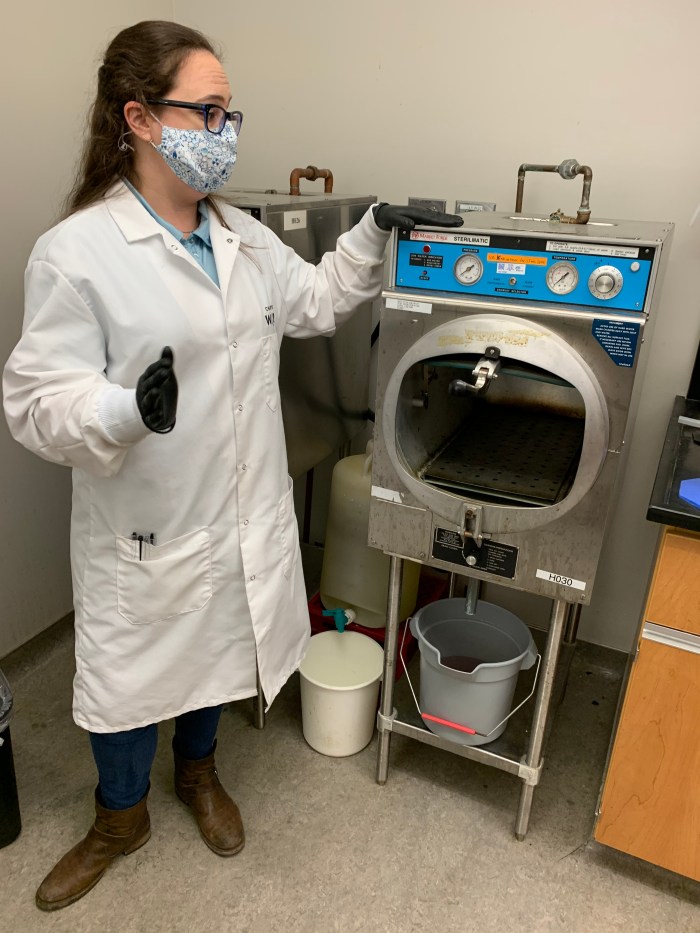lab technician wearing floral printed mask, glasses and long white lab coat stands next to tall, stainless steel testing device with blue trim and large round, black window with grey bucket underneath