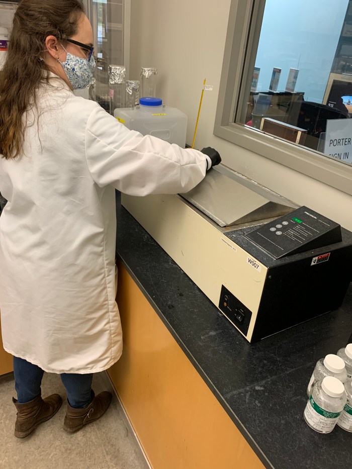 lab technician wearing glasses, floral printed mask and long white lab coat puts a grey cover on top of tan and black warming console