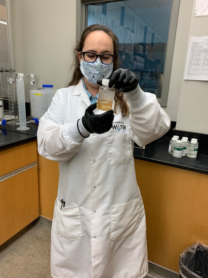 lab technician wearing glasses, floral print mask, black gloves, and long white lab coat examines the ginger-ale colored liquid inside a glass specimen jar with white lid