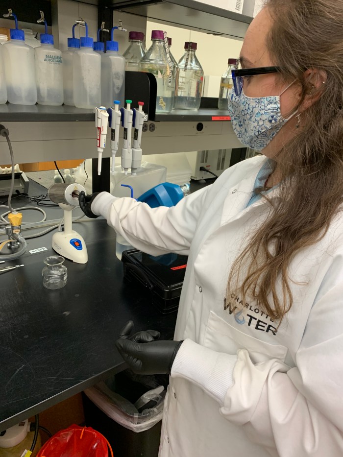 lab technician wearing glasses, floral print mask, black gloves and white lab coat with Charlotte Water logo, inserts a probe into a white testing device sitting on a black lab table
