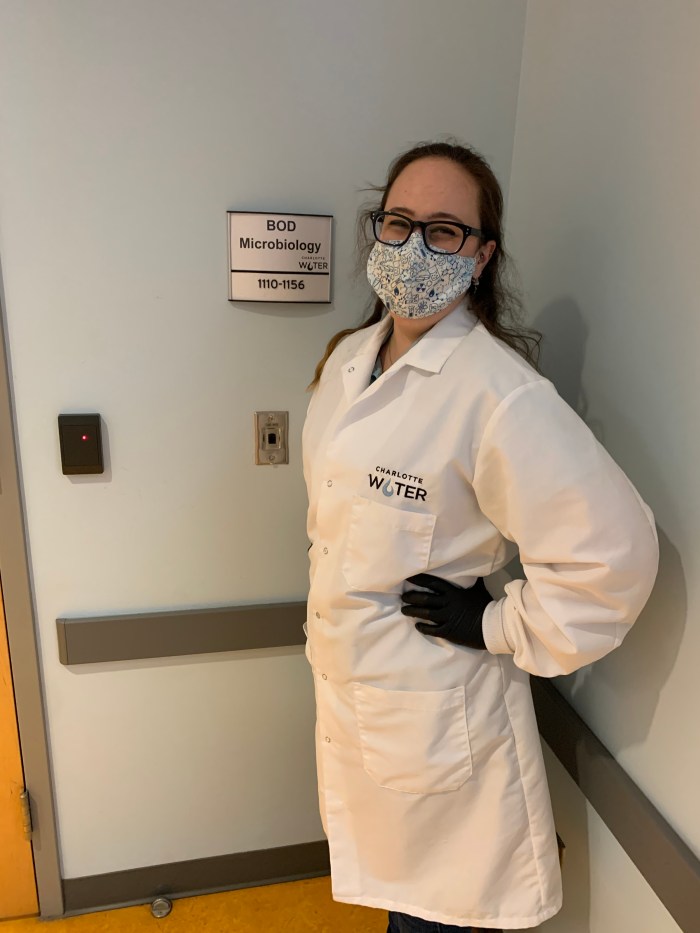 lab technician wearing glasses, floral printed mask, long white lab coat and black gloves poses for photo while standing in corner of elevator with light grey wall
