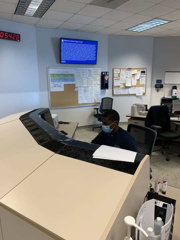 utility employee with blue mask sits behind a large tan and black console in an office with several bulletin boards and a large screen monitor on the walls behind him