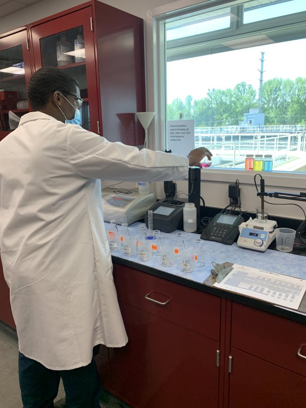 lab technician in white lab coat looks out lab window at water treatment facility treatment pools while sets of clear glass beakers and specimen jars sit on a blue and black lab table in front of him along with a clipboard with white papers