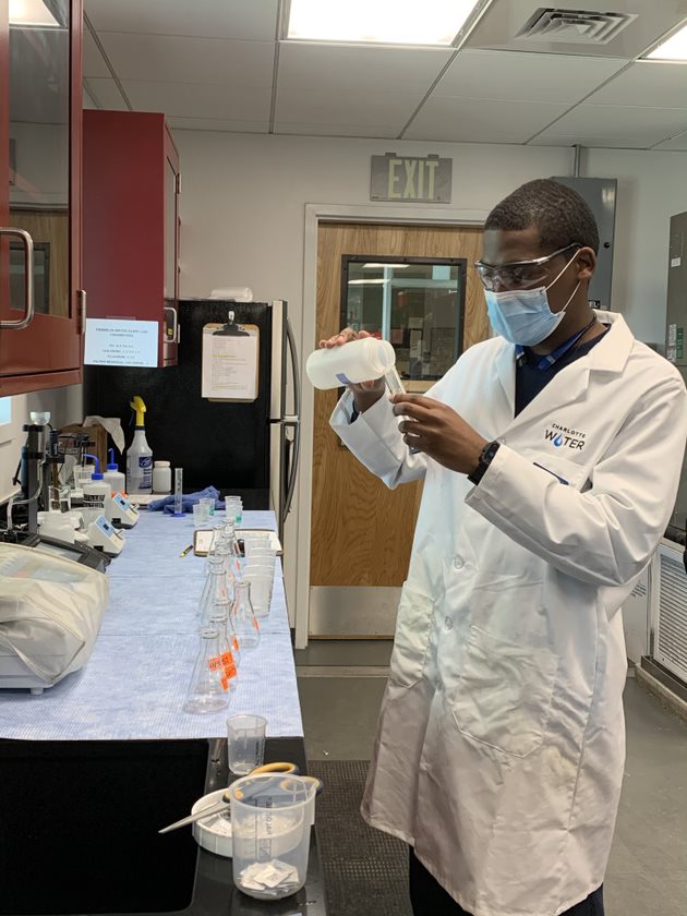 lab technician wearing long white lab coat, blue mask and glasses pours liquid out of an opaque plastic specimen jar into a clear glass test tube as he stands next to a blue lab table filled with various glass beakers