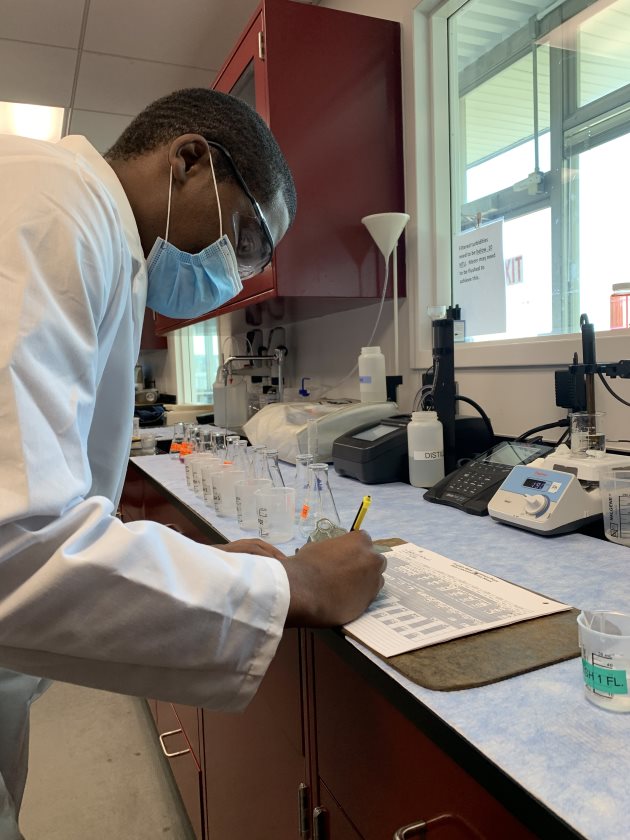 lab technician in white lab coat, blue mask and glasses bends over at light blue lab table to fill out a white paper form on a brown clipboard, nearby are a dozen glass beakers with companion small specimen jars