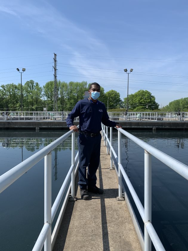utility employee in navy blue polo shirt and blue mask stands on a concrete catwalk with metal railings in between two large rectangular water treatment pools