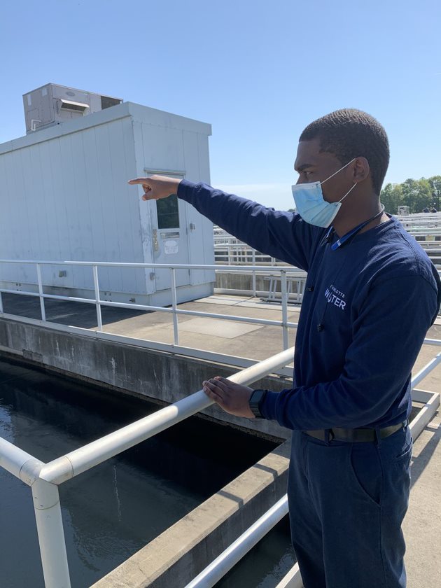 utility employee with navy blue polo shirt and blue mask stands next to a white railing, gesturing across the water treatment pool in front of him