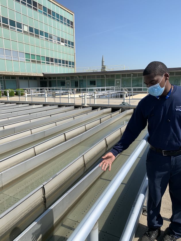 panoramic view of a utility employee in navy blue polo shirt and blue mask standing alongside a fenced in large treatment pool with nearly a dozen rectangular filter troughs filled with water, the employee gestures at one of them