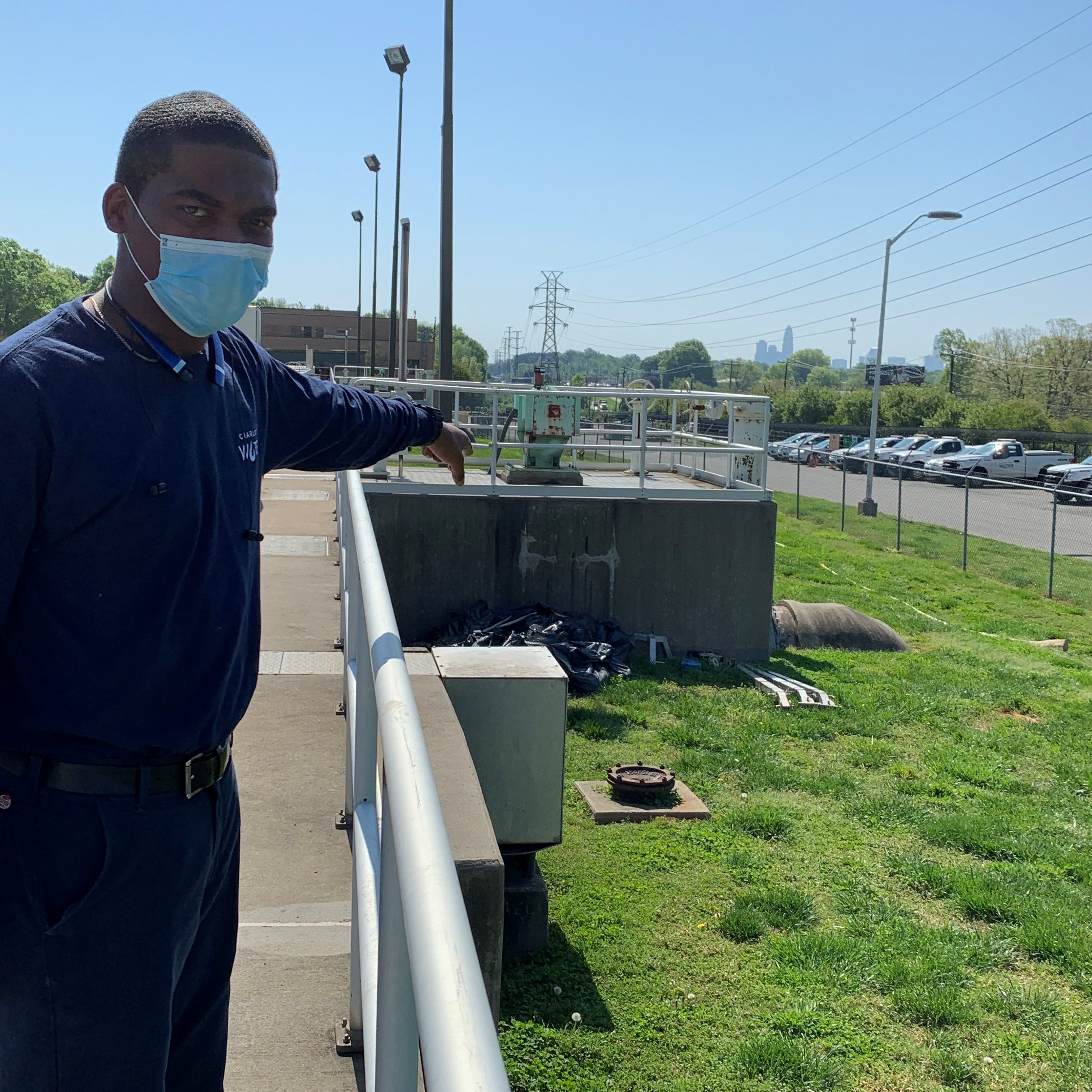 utility employee standing outside with blue mask points behind him at a large brown treatment plant water pipe emerging from ground connected to a green metal box installed atop a large concrete square platform