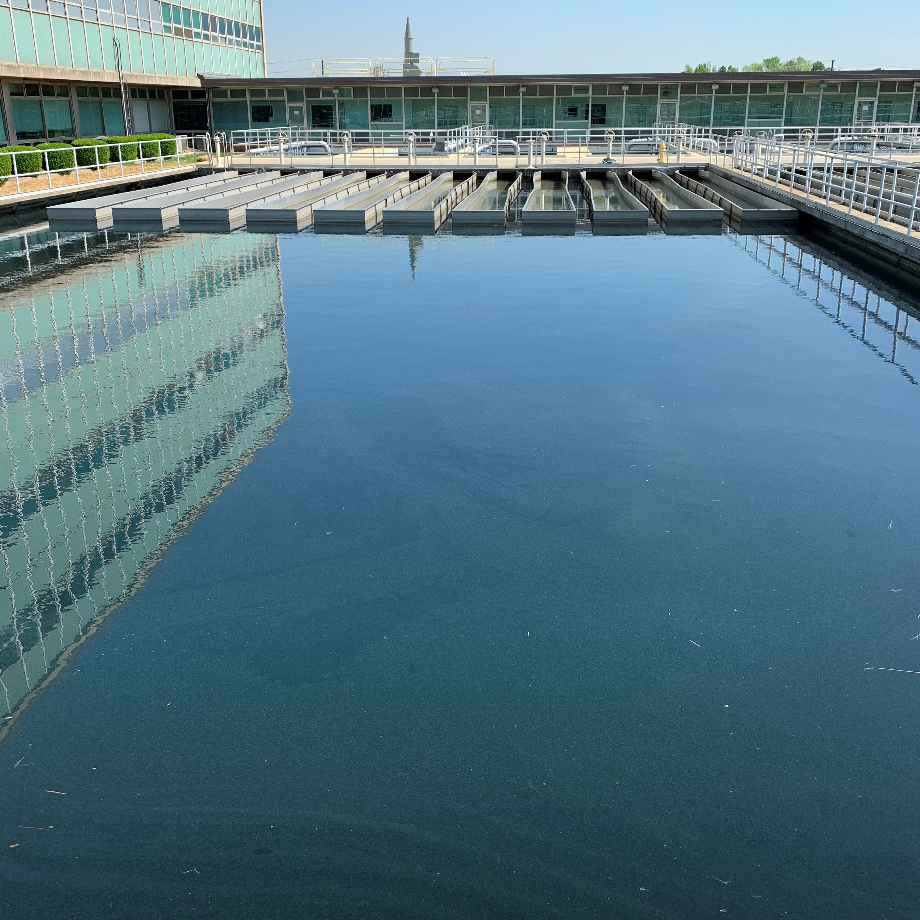 Panoramic view of water treatment plant's building next to large outdoor, rectangular sedimentation pond with row of 11 smaller rectangular filter troughs inside, metal fencing surrounds all water areas