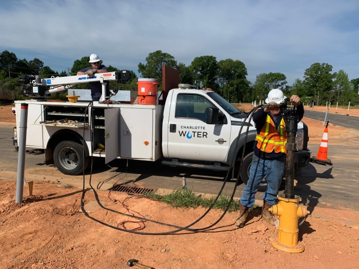 2 black pneumatic hoses run from a white utility truck with Charlotte Water logo into a tall brown pole-shaped tool held by a utility technician wearing orange safety vest and white hard hat as he holds the tool in place inside a new yellow fire hydrant he is installing roadside