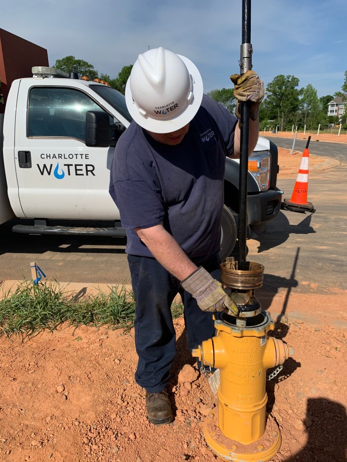 utility technician wearing navy blue t-shirt and white helmet stands next to newly installed yellow fire hydrant, he uses a black pole to lower a copper cufflink type valve inside of the hydrant's tower, a white utility truck with Charlotte Water logo on door an orange traffic cone appear behind him on the roadway