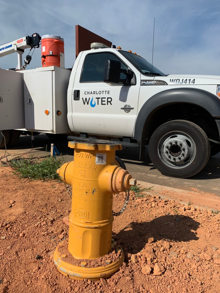 new yellow fire hydrant in process of being installed in recently dug reddish earth dirt awaits to have its cap bolted on as it sits curbside next to white utility truck with Charlotte Water logo on door