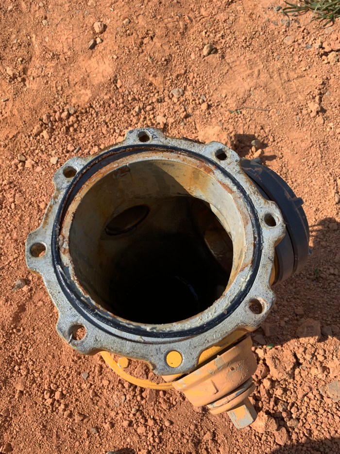 view looking down into a yellow fire hydrant without its top cover, the perimeter of the opening is a grey metal flange with holes for bolts, loose reddish brown dirt surrounds the unit