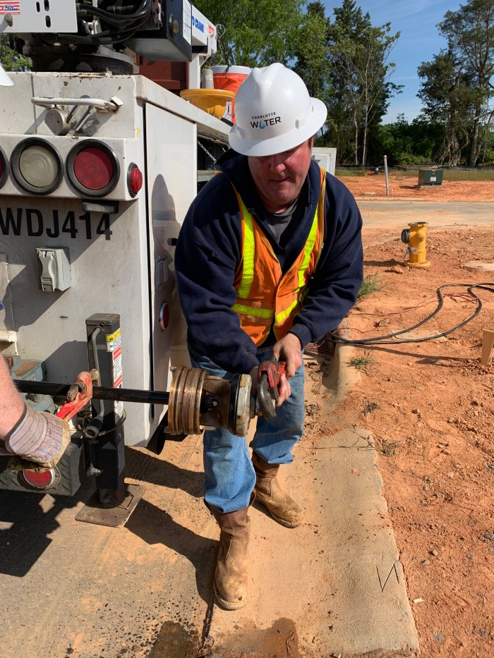 utility technician wearing orange safety vest and white hard hat uses an orange pipe wrench to grab hold of copper cuff-link topped part from its holder on the back of a white utility truck