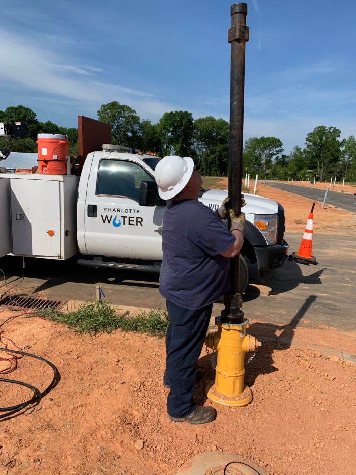 utility technician wearing navy blue t-shirt and white helmet stands next to newly installed yellow fire hydrant, he inserts a black pipe with a copper cufflink type valve inside of the hydrant's tower