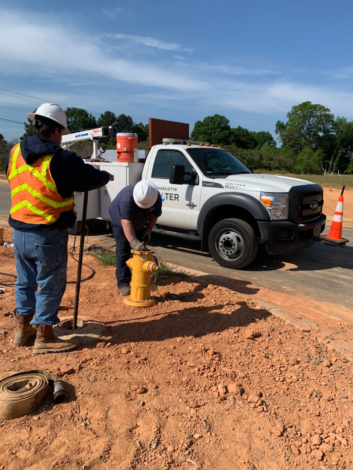 while utility technician in navy blue t-shirt and white hard hat bends down to work on the open top of a newly installed yellow fire hydrant, a second utility technician wearing an orange safety vest and white hard hat stands near by operating a tall metal key wrench he's inserted into a water meter hole in the dirt, also nearby is a white utility truck with Charlotte Water logo on door