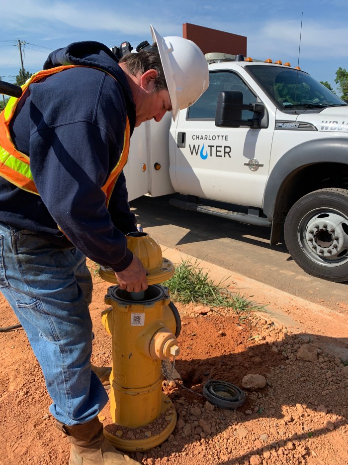 utility technician wearing orange safety vest and white hard hat bends over newly installed yellow fire hydrant as he places its yellow dome lid on top of its tower stack, a white utility truck with Charlotte Water logo is parked curbside nearby
