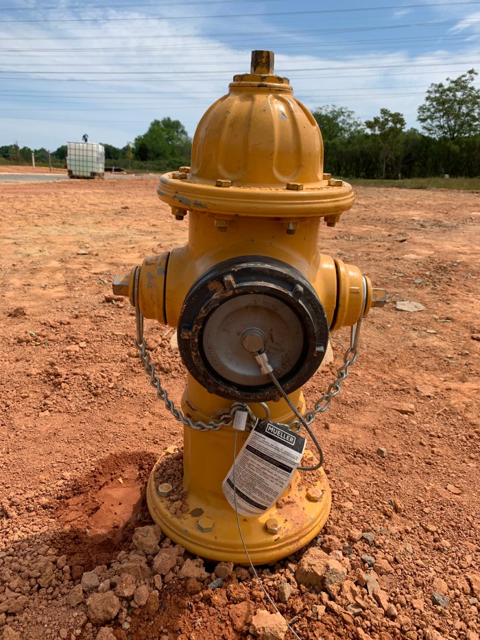 ground-level view of a newly installed yellow fire hydrant sitting in dirt, facing us with its black, round gasket front secured with a short cable and chains
