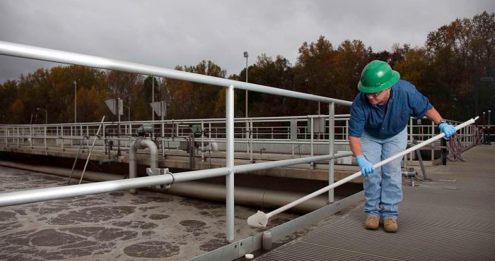water treatment plan technician in blue collared shirt and green hard hat bends down along metal catwalk next to a rectangular treatment pool of water to secure a water sample using a metal scoop on a long pole
