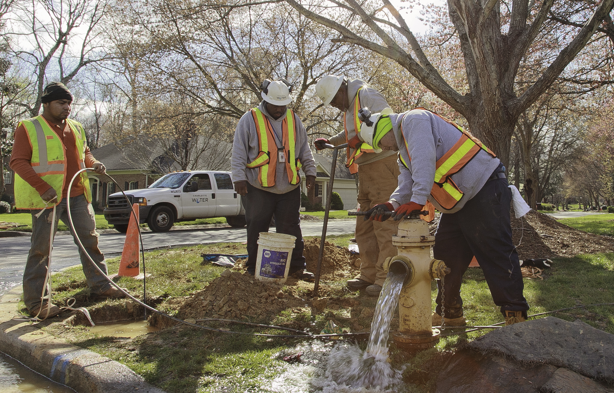 4 utility technicians wearing orange safety vests and white hard hats service a yellow fire hydrant on a street corner in a tree-lined suburban neighborhood, one man uses a pipe wrench to open the value of the hydrant causing water to pour out of its front while another man turns a tall pipe key inserted into a nearby water meter hole