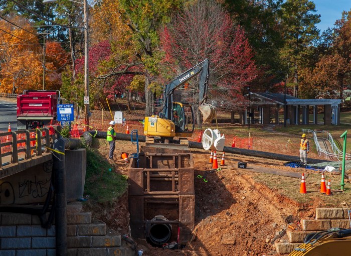 colorful autumn trees are the backdrop to a large black and yellow Deere excavator usiing a chain to prepare to lift a large white valve, in front as two orange-safety vest clad utility technicians with hard hats observe, in front is large, wide and deep trench with a stack of 3 rusty metal structures housing a large grey pipe coupler at bottom