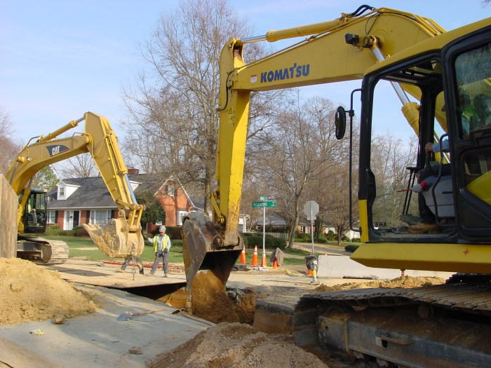yellow komatsu excavator remove reddish dirt from a trench dug on suburban street as 1 utility technician in yellow safety vest and white hard hat stands directing how a second yellow excavator using a chain removes a large brown steel plate covering a portion of the trench from the roadway
