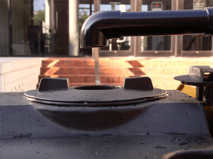 elbow of a black pipe points downwards to black flange and opening on a black tank beneath it