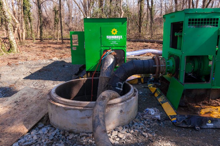 2 large green pumping devices parked on dirt in a forested area are attached to 2 black large hoses that are running down into a large, round concrete opening