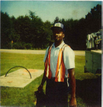 utility employee wears orang safety vest and with light blue short sleeved collared shirt, he stands next to large concrete base installed on grass