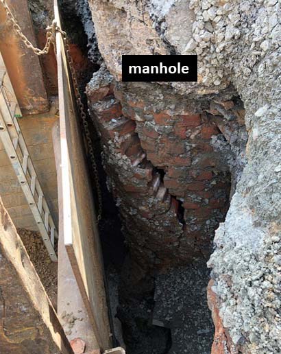 excavation of a trench reveals the side of a manhole's rounded, brick lined exterior peeking through grey dirt and gravel, a black notification with white font on the photo says, Manhole