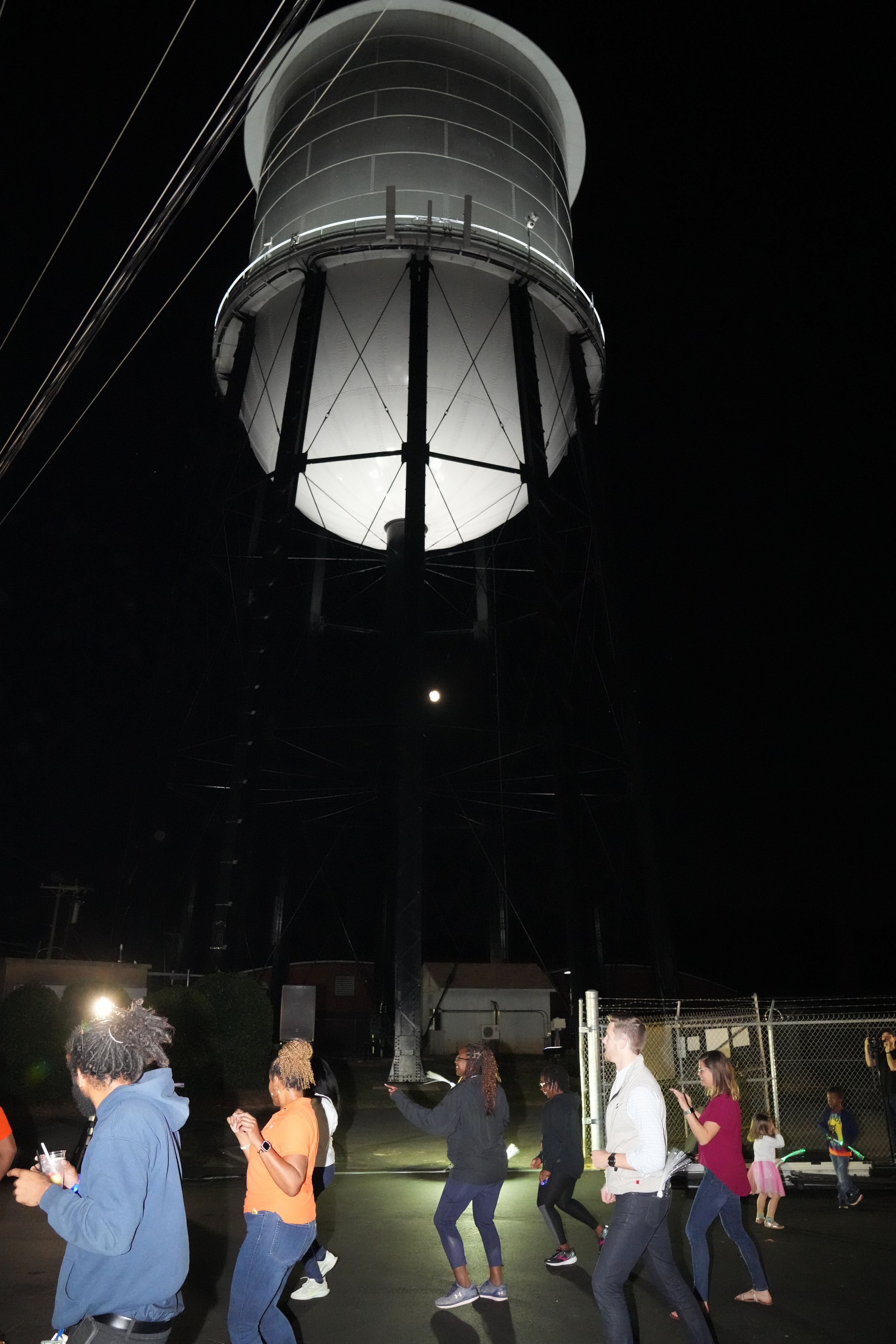 several utility employees in dance below the bulbous, illuminated white Charlotte water tower at night