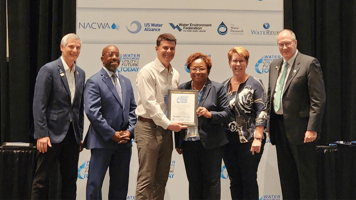 6 utility supervisors stand in front of a large water association sign for a group photo, woman in middle in dark suit along with man with cream shirt jointly hold a grey framed award plaque