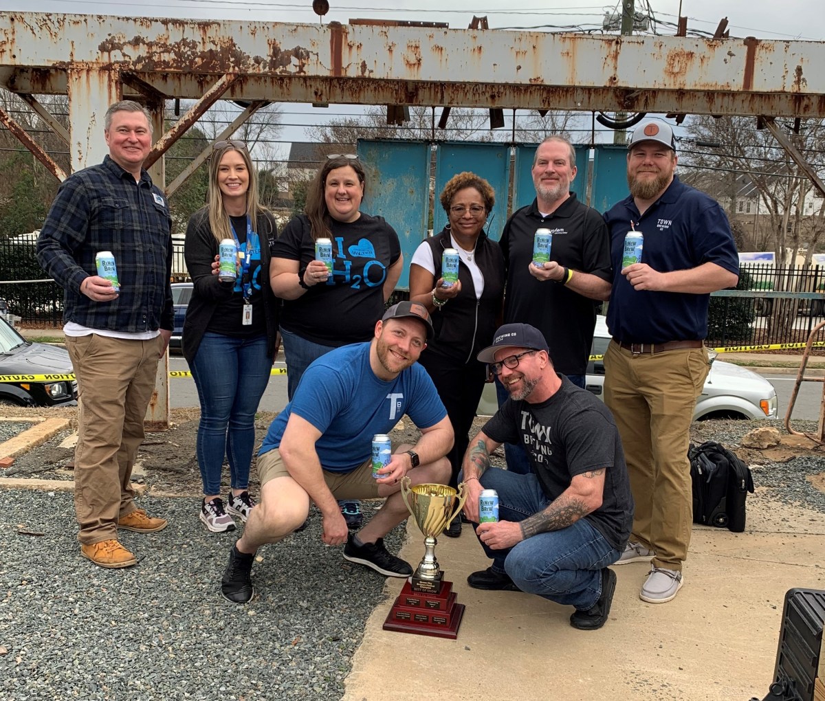 6 employees stand with 2 kneeling in front of them pose for a group photo with a large golden trophy, each person holds a light blue and green can of water